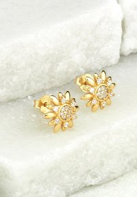 Orecchini a forma di fiore in oro, con un grappolo centrale di cristalli trasparenti e accenti a forma di petalo, su uno sfondo di pietra chiara.