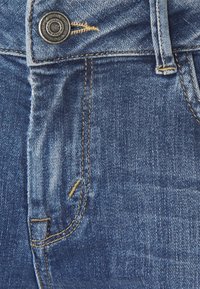 Denimjeans med en ljusblå tvätt, kontrasterande sömmar och en metallknapp. Texturen är något sliten nära fickorna.