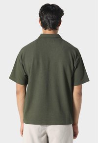 Lyhythihainen, oliivinvihreä paita, joka on valmistettu teksturoidusta kankaasta. Ominaisuuksiin kuuluu käännettävä kaulus ja rento istuvuus. Takapuolelta näkyy saumaton muotoilu.