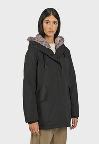 Černá parka s kapucí lemovanou umělým kožešinou, na zip a s druky, dvěma bočními kapsami a volným střihem, vyrobená z odolné textilie.