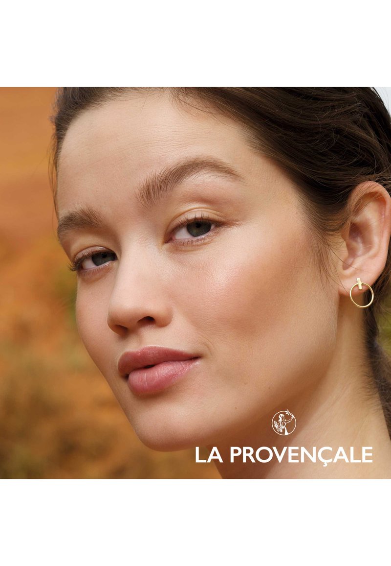 Boucle d'oreille en or sur un modèle au teint lisse et naturel. L'arrière-plan présente une tonalité terrestre chaude et floue. Texte : "LA PROVENÇALE."