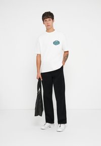 White short-sleeve t-shirt with a teal graphic on the chest, paired with black trousers and white sneakers. A black bag is held in one hand.