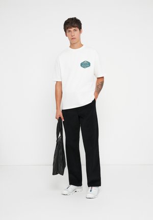 White short-sleeve t-shirt with a teal graphic on the chest, paired with black trousers and white sneakers. A black bag is held in one hand.