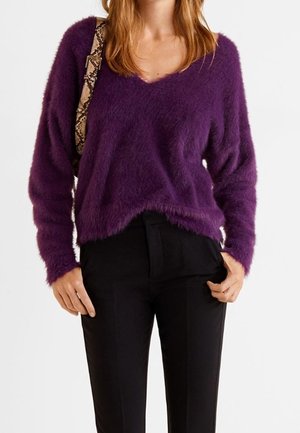 Kobieta w fioletowym, puszystym swetrze z dekoltem w serek, czarnych spodniach i torebce na ramię z paskiem w wzór skóry węża.