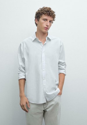 Light gray button-up shirt with long sleeves rolled to the elbows, soft texture, and a curved hem, paired with light beige pants.