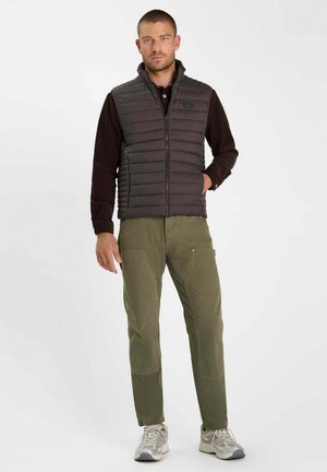 Uomo che indossa un gilet imbottito nero sopra una camicia nera a maniche lunghe, pantaloni verdi e scarpe da ginnastica grigie, in piedi davanti a uno sfondo semplice.