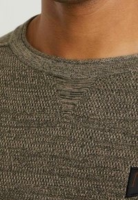 Pletený svetr v tlumené olivově zelené barvě s texturovaným vzorem. Kulatý výstřih, krátké rukávy a malá černá etiketa na lemu.