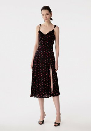 Donna che indossa un vestito midi nero con piccole stampe floreali rosse, spalline sottili con fiocchi e uno spacco alto sul lato sinistro, abbinato a tacchi neri.