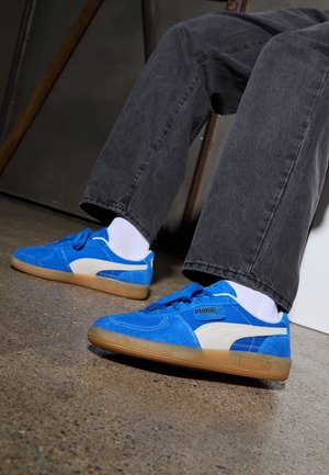 Personne portant des baskets en daim bleu Puma avec des bandes blanches, des chaussettes blanches et un jean gris foncé, debout sur un sol en béton poli.