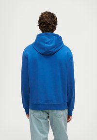 Μπλε βαμβακερό φούτερ με μεγάλη, ρυθμιζόμενη κουκούλα, φινιρίσματα σε ριμπ και στρίφωμα. Ο σχεδιασμός είναι casual, με ένα ενιαίο χρώμα χωρίς σχέδια.