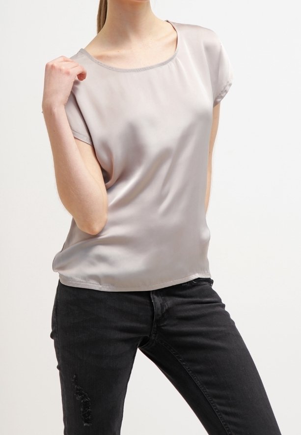 Femme portant un chemisier en satin gris clair à manches courtes et un jean noir déchiré, posant avec une main près de son épaule.