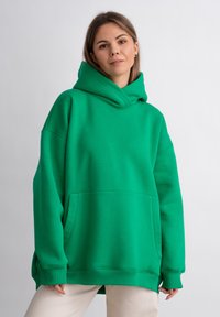 Felpa oversize verde con una grande tasca frontale, polsini a coste e un cappuccio spesso con stringa. Texture liscia con un design casual.