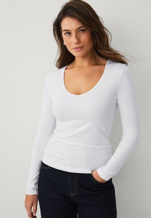 Frau mit langen braunen Haaren trägt ein weißes, langärmeliges Shirt mit Rundhalsausschnitt und dunkle Jeans, steht mit einer Hand in der Tasche vor hellem Hintergrund.