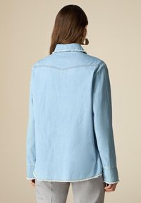 Light blue denim shirt with a relaxed fit, collar, and frayed hem. Features long sleeves and stitched detailing on shoulders and cuffs.
