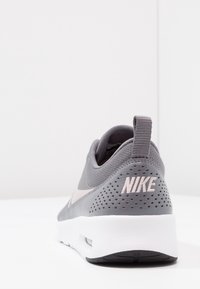 Šedá a bílá Nike teniska z pohledu zezadu vpravo, vybavená dvoucentimetrovým poutkem a perforovanou patou s jemně růžovým vyšitým logem.