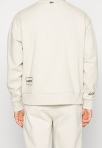 Personne portant un sweat-shirt et un pantalon de couleur crème, avec des logos Lacoste sur le haut du dos, le bas du dos et le poignet de la manche droite.