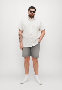 Cămașă cu mâneci scurte, de culoare bej deschis, cu dungi verticale, asortată cu pantaloni șort gri și adidași albi. Modelul poartă ochelari de soare.