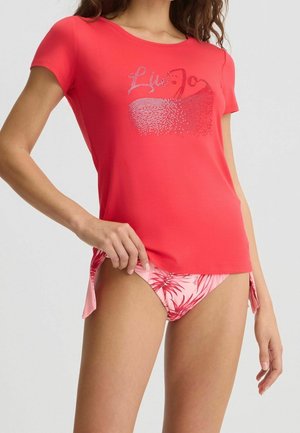 Frau trägt ein korallenfarbenes Kurzarmoberteil mit einem Schwan-Motiv aus Strasssteinen und pinke Bikinihöschen mit tropischem Muster und seitlichen Bindebändern.