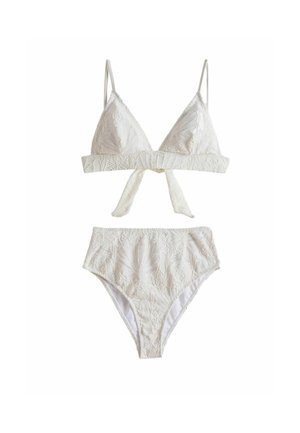 Jeune femme en bikini blanc taille haute debout sur une plage de sable avec une eau turquoise, regardant par-dessus son épaule.