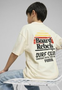 Camiseta de gran tamaño color crema con un gráfico retro en la parte posterior, que presenta "Board Rebels" y "Surf Club" en texto negro y naranja.