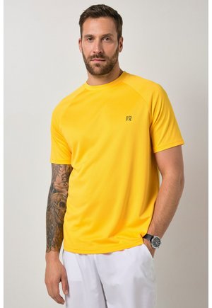 Mężczyzna o brązowych włosach i brodzie, ubrany w jasnżółtą krótką koszulkę z małym czarnym logo oraz białe spodnie, prezentujący wytatuowaną lewą rękę.