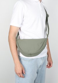 Persona che indossa una polo bianca e jeans azzurri chiari, con una borsa a tracolla verde oliva appesa sulla spalla, attraversando il torace.