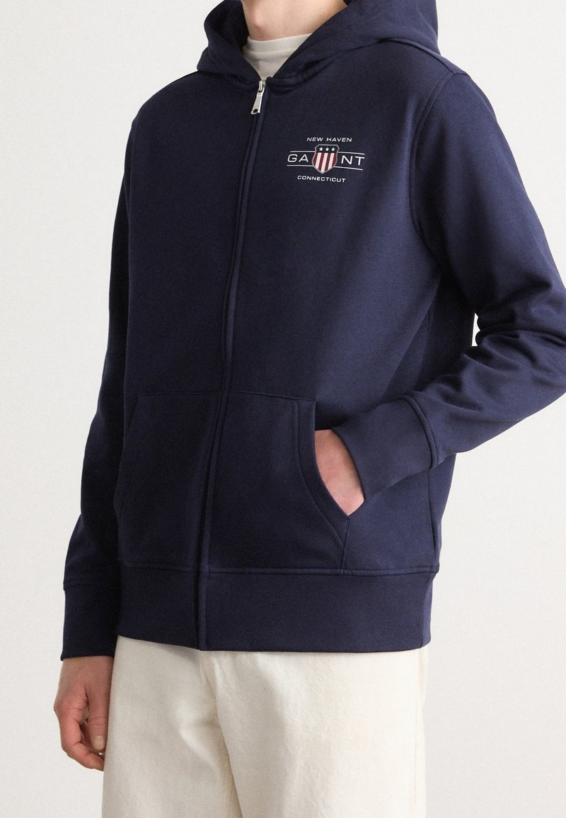 Haut à capuche zippé bleu marine en mélange de coton, avec une poche kangourou, un logo sur la poitrine et des poignets et un ourlet côtelés. Texture lisse.
