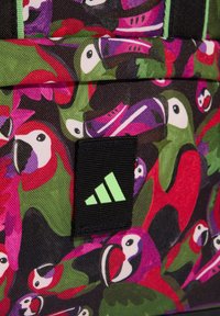 Zaino colorato con motivo a pappagallo, realizzato in tessuto resistente. Include una toppa nera con un logo verde. Superficie strutturata.