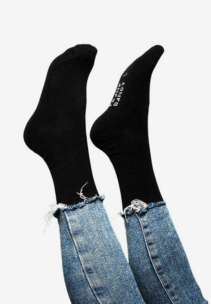 Schwarze Socken aus glattem Stoff, die an Füßen präsentiert werden, kombiniert mit ausgebleichten blauen Jeans, die ausgefranste Säume und eine eng anliegende Passform aufweisen.