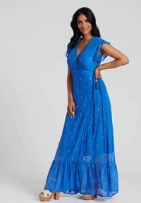 Abito lungo blu con maniche a volant corte, caratterizzato da un design a incrocio e motivi a diamante, realizzato in tessuto leggero. Sandali con piattaforma in legno.