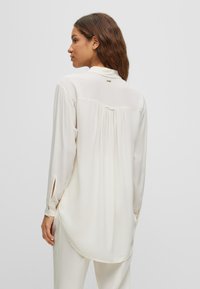 Balta ilgaa rankovė blouse su šilkinio audinio tekstūra, sagutėmis aptrauktomis rankovėmis, nugaros raukiniais ir mažo logotipo akcentu šalia apykaklės.