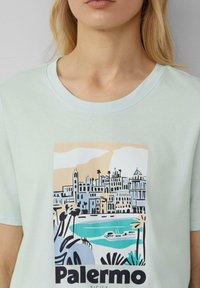 Licht mintgroene katoenen t-shirt met een grafische print van Palermo, Sicilië, met een gestileerde skyline, blauw water en zwarte tekst.