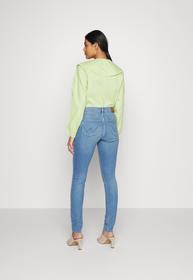 Krótką bluzkę w jasnym zielonym kolorze z dużym kołnierzem i długimi rękawami zestawiono z dopasowanymi, jasnoniebieskimi dżinsami oraz beżowymi klapkami na obcasie.