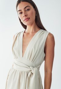 Robe enveloppante de couleur crème en tissu léger, avec un décolleté en V profond, des détails plissés aux épaules et une taille accentuée par un nœud.