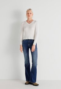 Balts adījuma kabeļu džemperis ar V veida kakla izgriezumu, apvienots ar platajiem tumšajiem džinsiem. Iestatīts neitrālā fonā, uzsvars uz apģērba tekstūru.