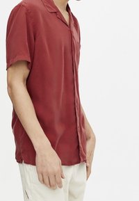 Chemise à manches courtes avec col, en tissu rouge pâle, dotée d'une patte de boutonnage et d'une coupe ajustée. Texture lisse avec une légère brillance.