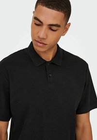 Sort polo t-shirt lavet af struktureret stof, med en klassisk krave og tre-knaps lukning. Ærmerne er korte og afslappede.