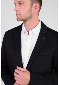 Zwarte blazer met notch lapel en enkele borstzak, gedragen over een wit overhemd met knopen, met een klein rond logo accent.
