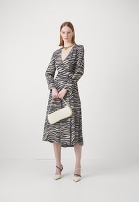 Beige ja tummansininen viidakko-kuvioinen mekko, jossa on pitkät hihat, syvä V-kaula-aukko ja vyötäröä korostava muotoilu. Yhdistettynä valkoiseen käsilaukkuun ja korkokenkiin.