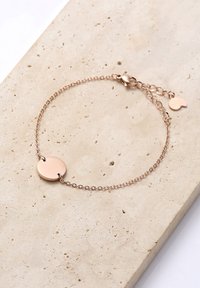 Rose gold bracelet with a round disc charm, featuring a delicate chain and adjustable clasp on a textured neutral stone surface.