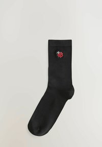 Zwarte sok van een glad materiaal, met een rode en zwarte lieveheersbeestje borduursel dicht bij de bovenkant. Verstelbare breedte en enkellengte.