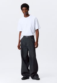 Bijela majica kratkih rukava uparenja s tamnim, širokim trapericama. Značajni detalji uključuju tamno šivanje i vidljivu kopču na dugme.