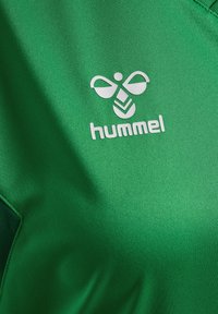 Logo blanc Hummel avec une abeille stylisée au-dessus du nom de la marque sur un tissu vert vif, montrant la texture et les détails des coutures.