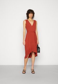 Vestido envolvente na cor ferrugem com decote em V profundo, cinto de laço na cintura e bainha assimétrica com babados. Usado com sapatos pretos de tiras e uma bolsa pequena.