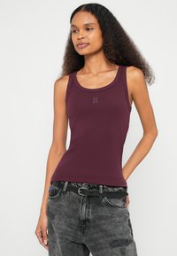 Viininpunainen ribbineuloksesta valmistettu hihaton paita, jossa on pyöreä pääntie ja pieni kohokuvioitu "HUGO"-logo rinnassa. Yhdistetty harmaiden farkkujen kanssa.