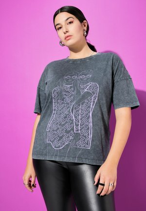 Una modelo posa sobre un fondo rosa, con una camiseta gris antracita con una ilustración de corazones y unos leggings negros brillantes.
