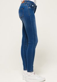 Vaqueros ajustados de mezclilla en color azul, que presentan una cintura de altura media, bolsillos traseros y un dobladillo limpio. Combinados con zapatillas blancas.