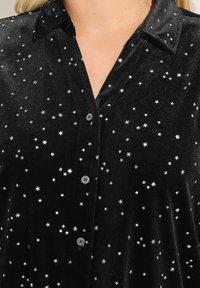Camicia in velluto nero con motivi a stelle argento, design con colletto, chiusura frontale a bottoni e texture morbida. I bottoni sono rotondi e scuri.