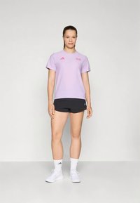 Noor naine seisab, seljas helesinine sportlik T-särk, mustad lühikesed püksid, valged sokid ja valged tossud, taust on ühtlane.