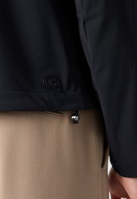 Black jacket with smooth texture, featuring a round logo and an adjustable drawstring at the waist, paired with beige textured trousers.